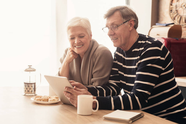 Casal idoso alegre usando tablet digital na cozinha juntos, lendo notícias
 - Foto, Imagem
