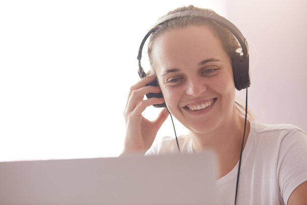 Smiling happy woman with headphones on head, sitting at desk in front of opened portable computer, looks at laptop screen, having video call. Pretty woman working on her notebook with headphones on. - Φωτογραφία, εικόνα