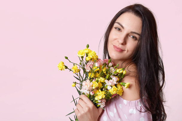 Indoor shot of young adorable brunette woman with bouquet of yellow flowers, gentle girl wearing pink sundresses with bare shoulders, model looking directly at camera, isolated over rosy wall. - Φωτογραφία, εικόνα