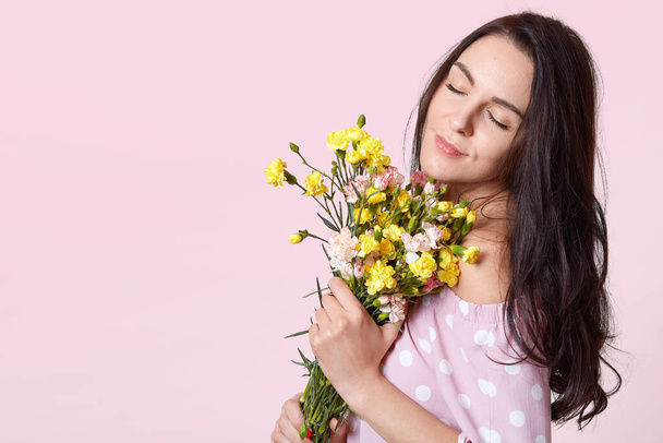 Close up portrait of beautiful young girl with bouquet of flowers, adorable dark haired lady with closed eyes holding flowers, gentle girl. Copy space for your advertisment or promotional text. - Foto, afbeelding