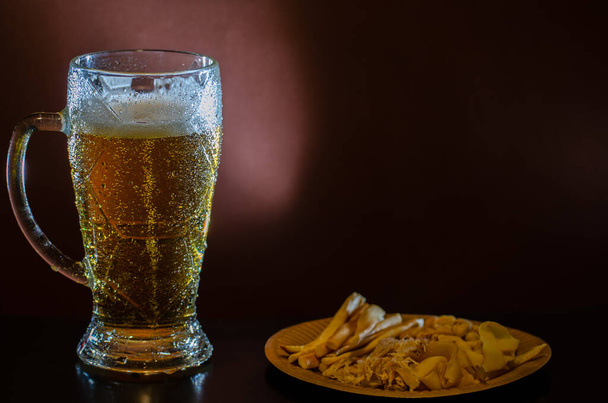 Ένα θολό ποτήρι ελαφριάς μπύρας με ένα σνακ σε σκούρο φόντο.. Κοντινό πλάνο, μακέτα. - Φωτογραφία, εικόνα