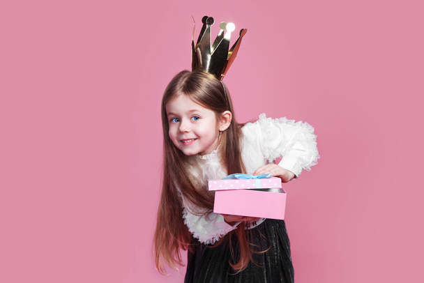 surprised little girl in crown and a white dress holding birthday present on pink background - Foto, afbeelding