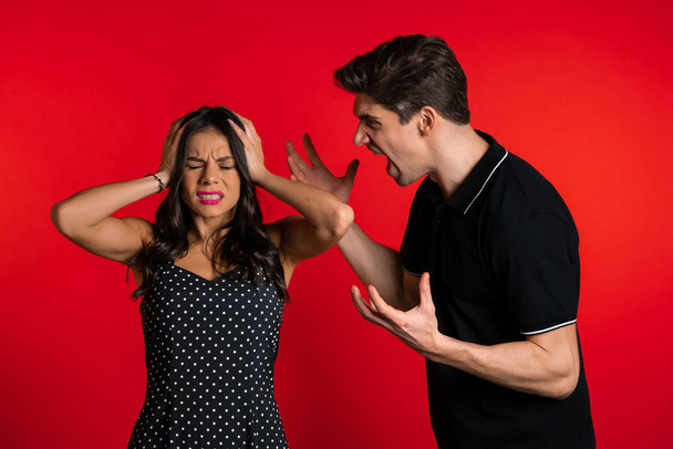 Young man emotionally screaming at his wife or girlfriend on red background in studio. Bored woman covers ears with hands. Concept of conflict, problems in relationships. - Фото, изображение