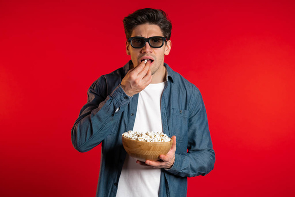 Young european man in 3d glasses watching fascinating comedy movie, laughing and eating popcorn on red studio background. - Фото, изображение