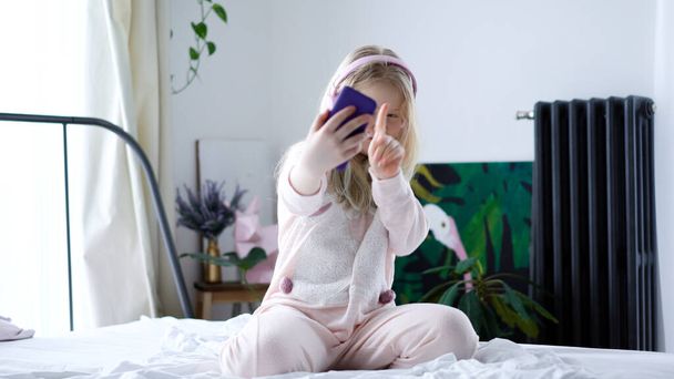 vida moderna de la generación Z. adolescente en pijama y auriculares en la habitación en la cama escucha música desde un teléfono inteligente
. - Foto, Imagen