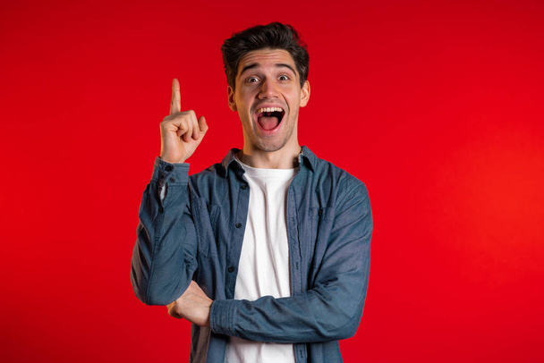 Portrait of young thinking pondering man having idea moment pointing finger up on red studio background. Smiling happy guy showing eureka gesture. - Фото, зображення