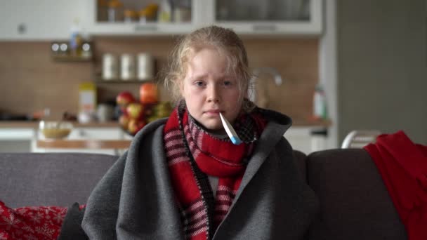 kaukasisches Mädchen zu Hause. Lustiges Porträt eines kranken Kindes in Schal und Karo mit Thermometer auf dem Sofa in der Wohnung - Filmmaterial, Video