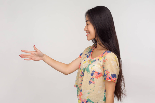Vista lateral de chica asiática amigable con pelo largo morena en blusa dando la mano al apretón de manos y sonriendo al cliente, saludando y conociéndose. estudio de interior tiro aislado sobre fondo gris
 - Foto, Imagen