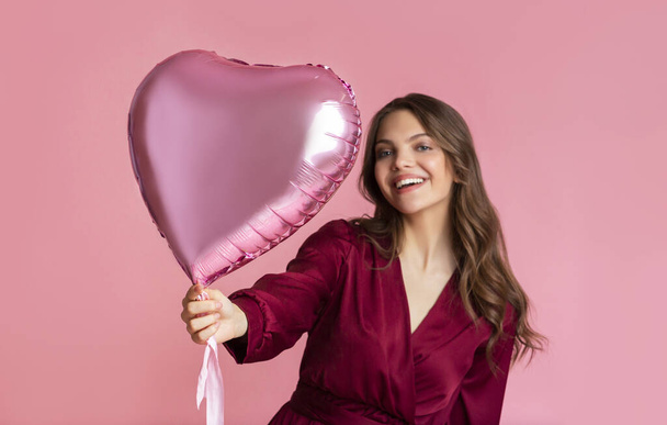 Heart-shaped balloon in hand of beautiful young woman over pink background - Zdjęcie, obraz