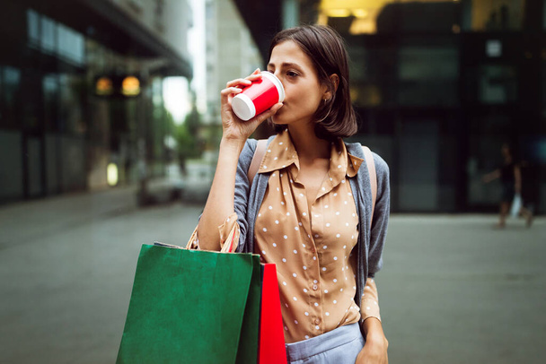 Happy young beautiful woman drinking take away coffee and walking with bags after shopping in city. - Foto, Imagem