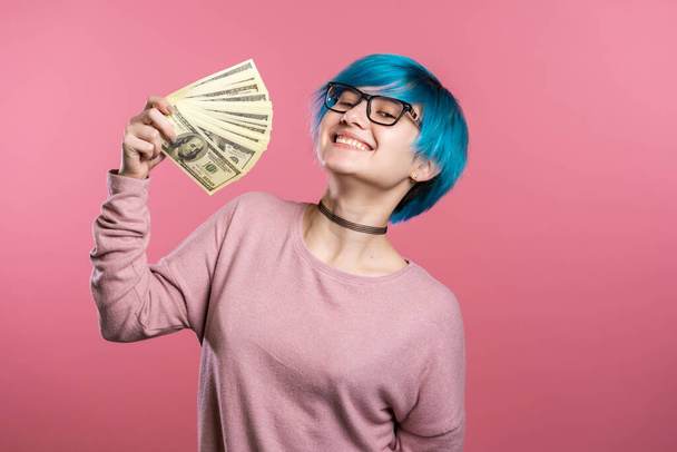 Satisfied cool excited girl with blue hair showing money - U.S. currency dollars banknotes on pink wall. Symbol of success, gain, victory. - Fotoğraf, Görsel
