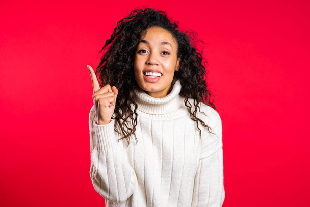 Portrait of young thinking pondering afro woman having idea moment pointing finger up on red studio background. Smiling happy girl showing eureka gesture - Fotografie, Obrázek