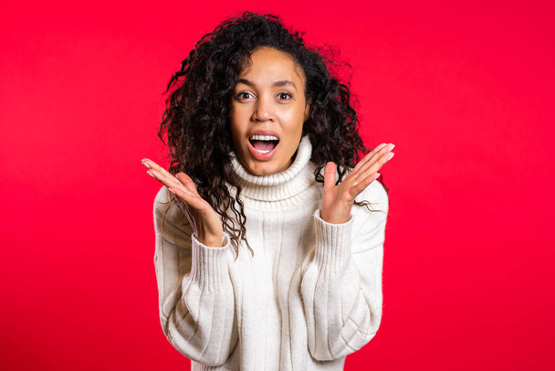 Pretty female shocked model on red background. OMG, wow effect. African girl with long hair with delight. Surprised excited happy woman in sweater - Valokuva, kuva