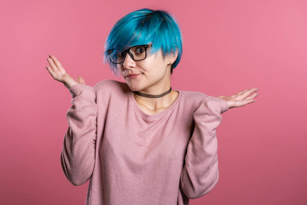 Young unsure girl with unusual appearance shrugs her arms, makes gesture of I dont know, care, cant help anything .Young millenial woman with blue hair on pink background - Photo, Image