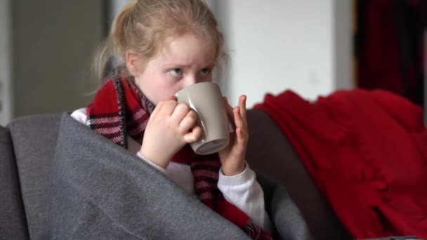 portret chorego dziecka w szaliku i kratę z filiżanką gorącej herbaty na kanapie w mieszkaniu - Materiał filmowy, wideo