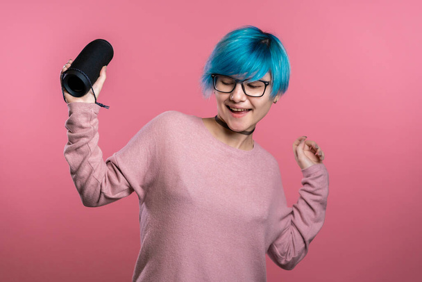 Cute woman with blue hair smiling and dancing with wireless portable speaker in pink studio background. Music, dance concept - Valokuva, kuva