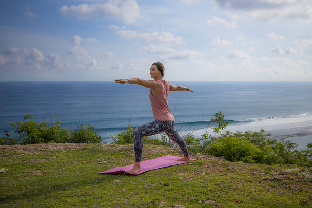Yoga yapan genç bir kadın, Virabhadrasana pozunda duruyor. Uçurumda açık hava yogası. Savaşçı pozu. Çalış. Sağlıklı bir yaşam tarzı. Arka planda okyanus. Yoga geri çekilme, Bali. - Fotoğraf, Görsel