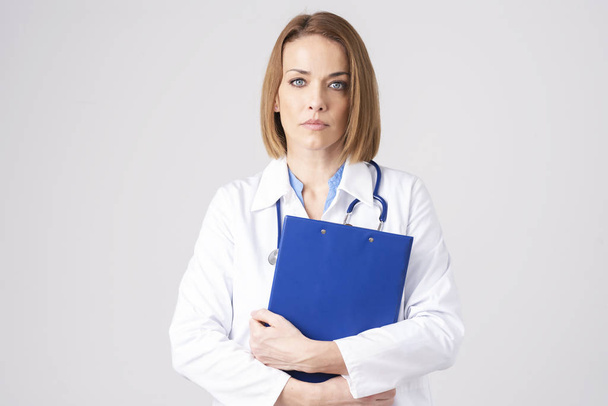 Portrait shot of middle aged female doctor wearing medical uniform and holding clipboard in her hand while standing at isolated white background. - Photo, Image
