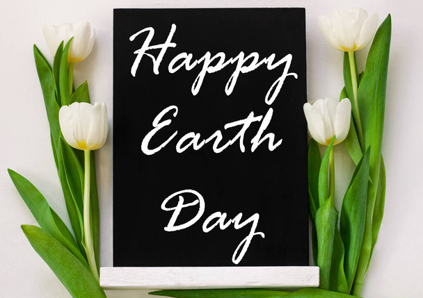 Feliz Día de la Tierra 22 de abril signo de saludo en pizarra negra con flores de tulipán sobre fondo blanco. World ecology concept card.Saving ambiente, guardar planet.Blackboard limpio con texto, pancarta
 - Foto, Imagen