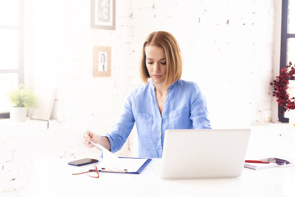 Tiro de atraente mulher de negócios madura fazendo alguma papelada enquanto sentada atrás de seu caderno na mesa de escritório
. - Foto, Imagem