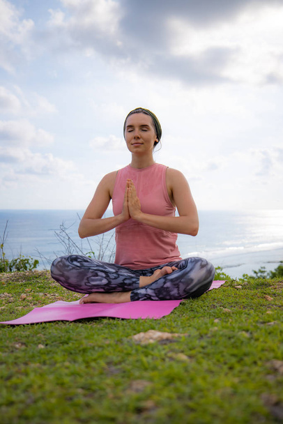 Young Caucasian woman sitting on yoga mat, meditating, practicing yoga and pranayama. Hands in namaste mudra. Outdoor yoga on the cliff. Zen lifestyle. Bali, Indonesia - Photo, Image