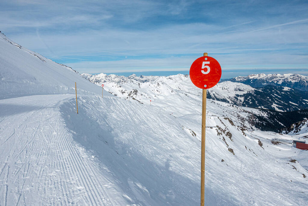 a red sign with the number 5 stands next to a snow covered ski route and the sky is blue - 写真・画像