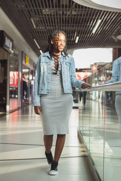 Молодая темнокожая женщина, одетая в модную одежду, позирует в торговом центре, размытый фон
 - Фото, изображение