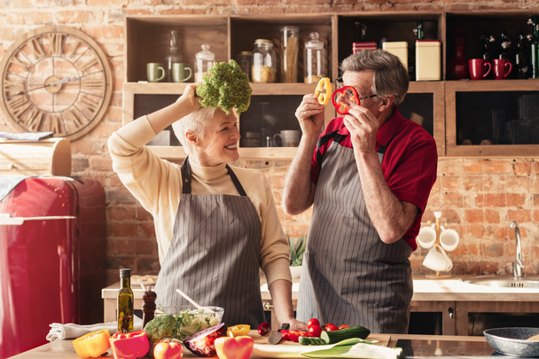 Elderly couple having fun in kitchen, playing with vegetables - Fotó, kép