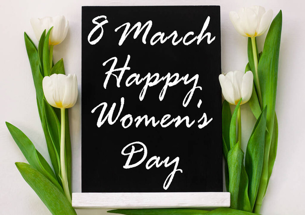 Happy Women's Day 8 March sign on black chalkboard with tulip flowers on white background. International Womens Day message written on blackboard display top view.Greeting card concept,flat lay banner - Photo, Image