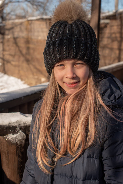 Porträt einer schönen Blondine mit Hut vor dem Hintergrund der Stadtlandschaft in der kalten Jahreszeit. Mädchen, das die Straße hinuntergeht. Mädchen mit schwarzem Hut und blauer Daunenjacke im Winter. Mädchen 9 Jahre alt. - Foto, Bild