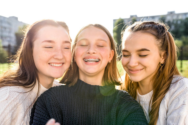 Retrato de cerca, tres niñas colegialas adolescentes, verano al aire libre. Feliz juego sonriente, teléfono de fotografía, foto selfie. Emociones felicidad diversión sonrisa, relajación deleite placer. Frenos en los dientes
. - Foto, imagen