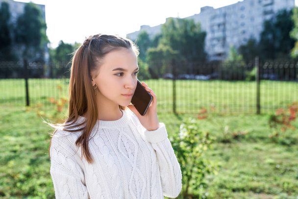 Teen girl in white sweater in summer in city. Calls on smartphone, listens to voice message. Casual wear. Free space for copy text. Green grass lawn building background. - Foto, Imagem