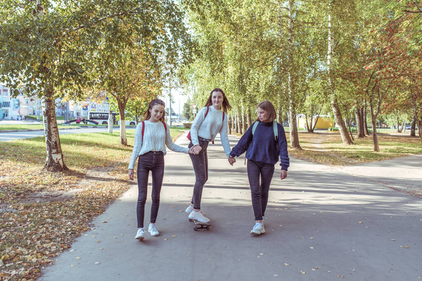 Three schoolgirls girls girlfriends in fall in city, ride a skateboard, in summer in park, return from school and college, behind back of bag are backpacks. Casual wear, sweaters, jeans, sneakers. - Fotografie, Obrázek
