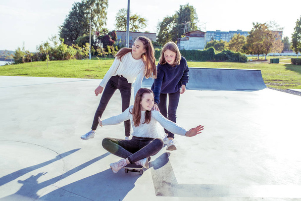 Young girls teenage schoolgirls ride skateboard summer park. Casual wear sweater jeans. Emotions happiness, fun, relaxation, smile laughter, best girlfriends, rest after college, free space for text. - Foto, imagen