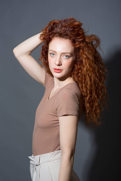 Poważna, rudowłosa, kręcona kobieta, patrząca prosto w kamerę, trzyma włosy w dłoni. Na odizolowanym szarym tle - Zdjęcie, obraz