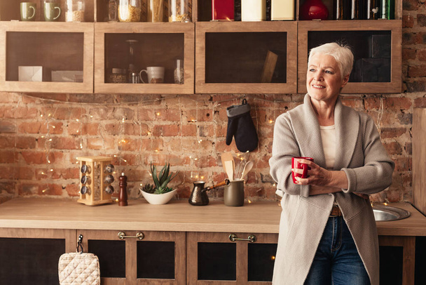 Mulher idosa relaxada bebendo café na cozinha e olhando para o lado
 - Foto, Imagem