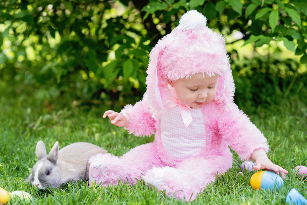  庭で草の上に描かれたイースターエッグに座っている間、ウサギと遊ぶウサギの衣装を着た小さな幼児の女の子。休日のコンセプト  - 写真・画像