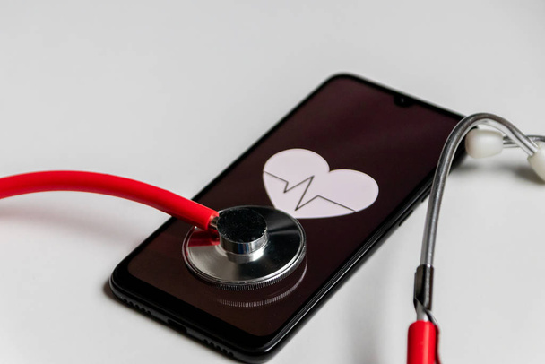 Stethoscope και μαύρο smartphone με ένα σύμβολο καρδιάς και καρδιακό παλμό για να δείξει ψηφιακό γραφείο γιατρούς, ψηφιακή διάγνωση και tracker υγείας, καθώς και στατιστικές υγείας και φάρμακα βοηθοί υγείας - Φωτογραφία, εικόνα
