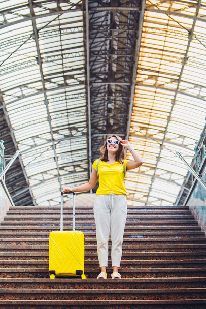 χαμογελαστή ευτυχισμένη γυναίκα με κίτρινη βαλίτσα σε ρόδες στις σκάλες του σιδηροδρομικού σταθμού αντιγράψτε την έννοια διαστημικό ταξίδι - Φωτογραφία, εικόνα