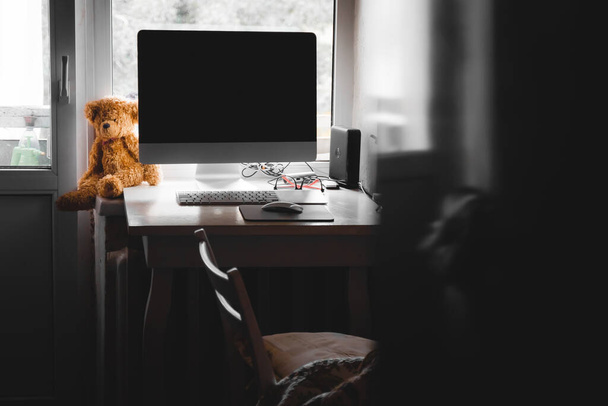 In het thuiskantoor zit een gele beer pluche speeltje op een tafel naast een computer monitor. Waas, selectieve focus, kopieerruimte. - Foto, afbeelding