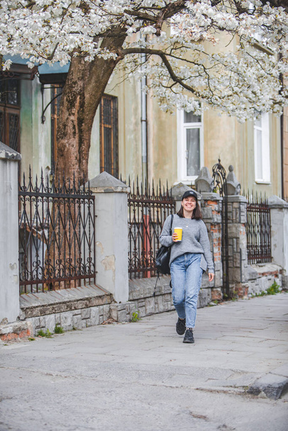 young stylish woman walking by street with coffee cup. spring blooming tree on background - Photo, Image