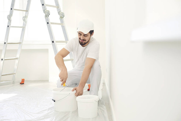 A male painter in a white uniform with a roller works in his hand in a white room - Fotoğraf, Görsel