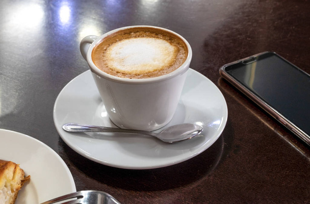 Bir fincan cappuccino, bir kafede latte, bir masanın üzerinde, siyah ahşap bir yüzey, akıllı telefonun yanında. Randevu, bekleme konsepti. Konsept kafede kahvaltı. Yatay yönelim. - Fotoğraf, Görsel