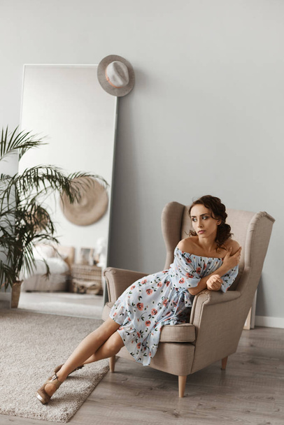 Beautiful young woman in a light silk dress with floral pattern sitting in armchair. Model girl with curly hair and slim body in light spring dress posing in the interior. Spring fashion - Photo, image