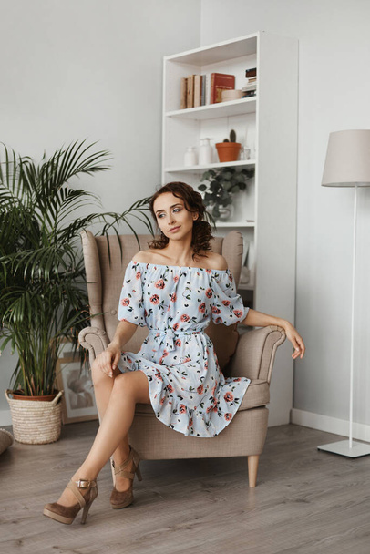 Fashion shot of a young beautiful woman in a short dress sitting in armchair. Portrait of a model girl with curly hair and slim body in light spring dress in the interior. Spring fashion. Summertime - Foto, imagen