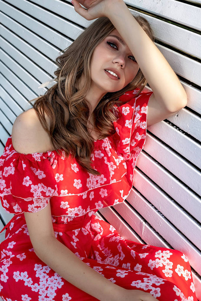 Beautiful young girl, model, brown-haired woman with long hair in a red dress sits on a bench and poses with a relaxed, languid look. Background white bench in the park - Foto, Imagem