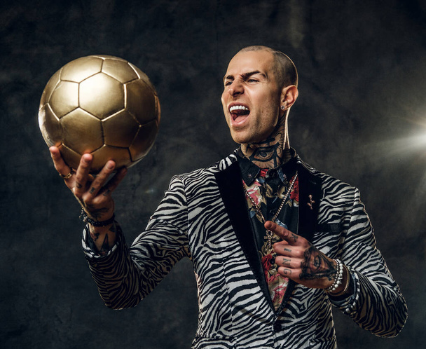 Bold tattooed male model posing for photoshoot in a dark studio and holding a golden soccer ball while screaming on it - Photo, Image