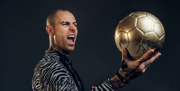 Смелый татуированный мужчина-модель позирует для фотосессии в темной студии и держит золотой футбольный мяч, крича на нем
 - Фото, изображение