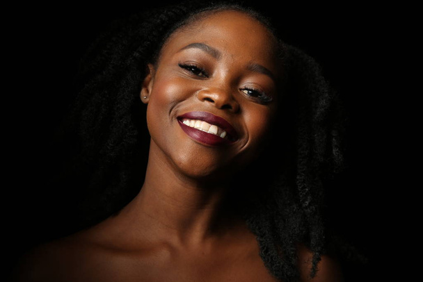 Porträt einer jungen schönen afrikanischen Frau mit dicken schwarzen Locken auf einem isolierten schwarzen Hintergrund. Die Frau lächelt - Foto, Bild