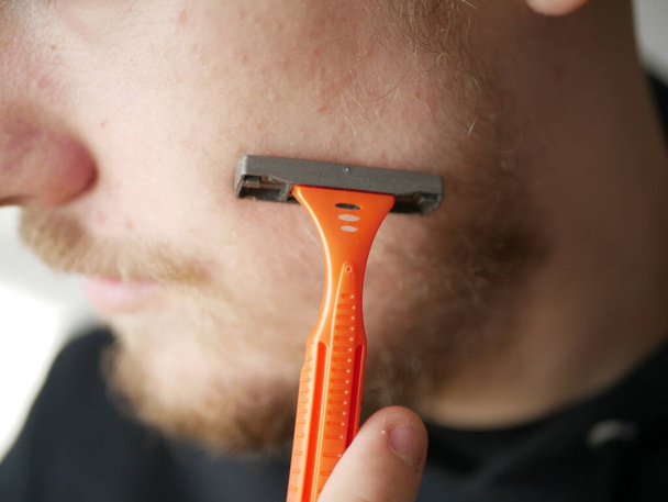 Jeune homme gars avec barbe montrant lame de rasoir orange jetable, studio tourné sur fond blanc
. - Photo, image